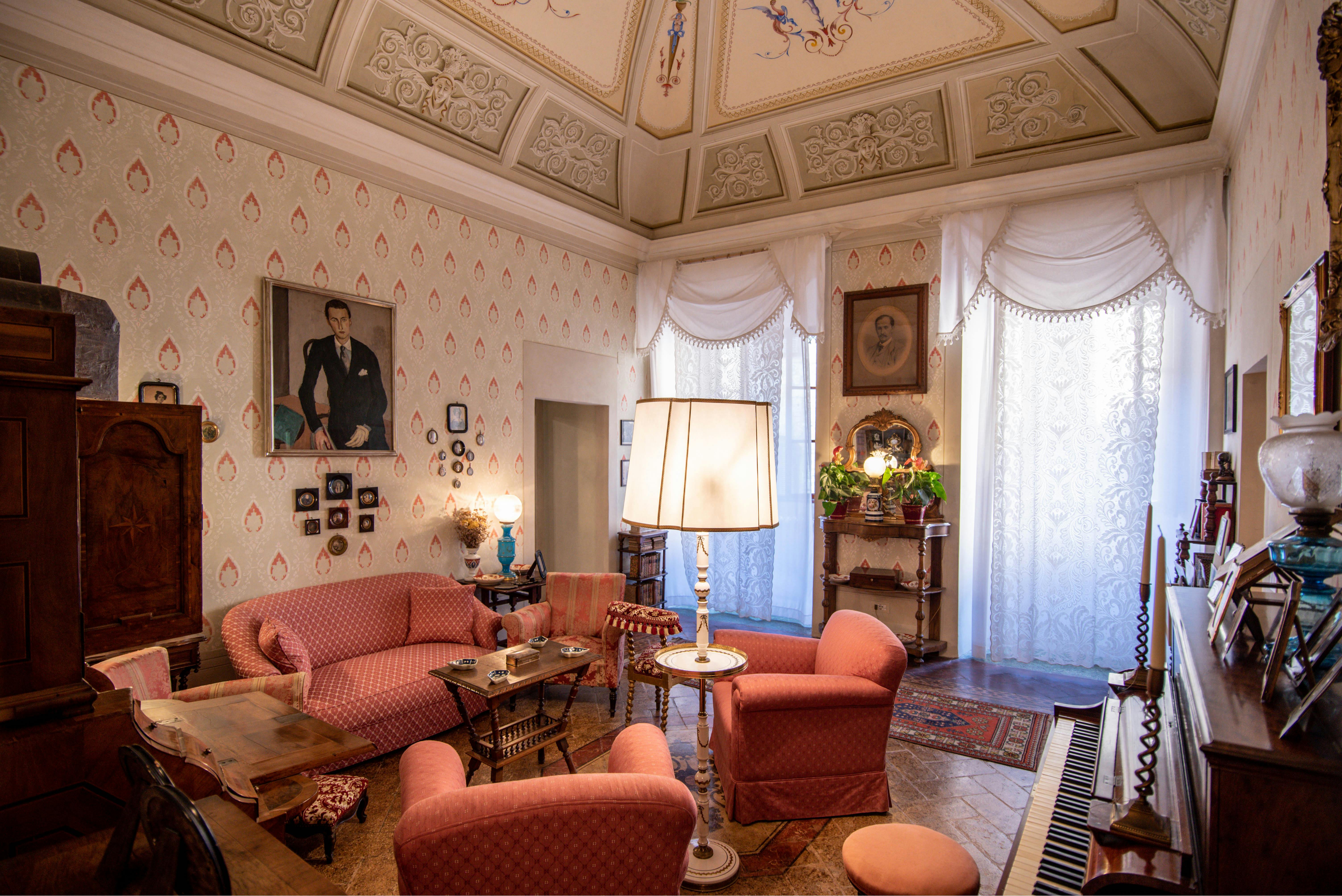 Un soggiorno vintage con carta da parati a fantasia, mobili rossi, soffitto decorato, grande lampada da terra, ritratti incorniciati e tende di pizzo.