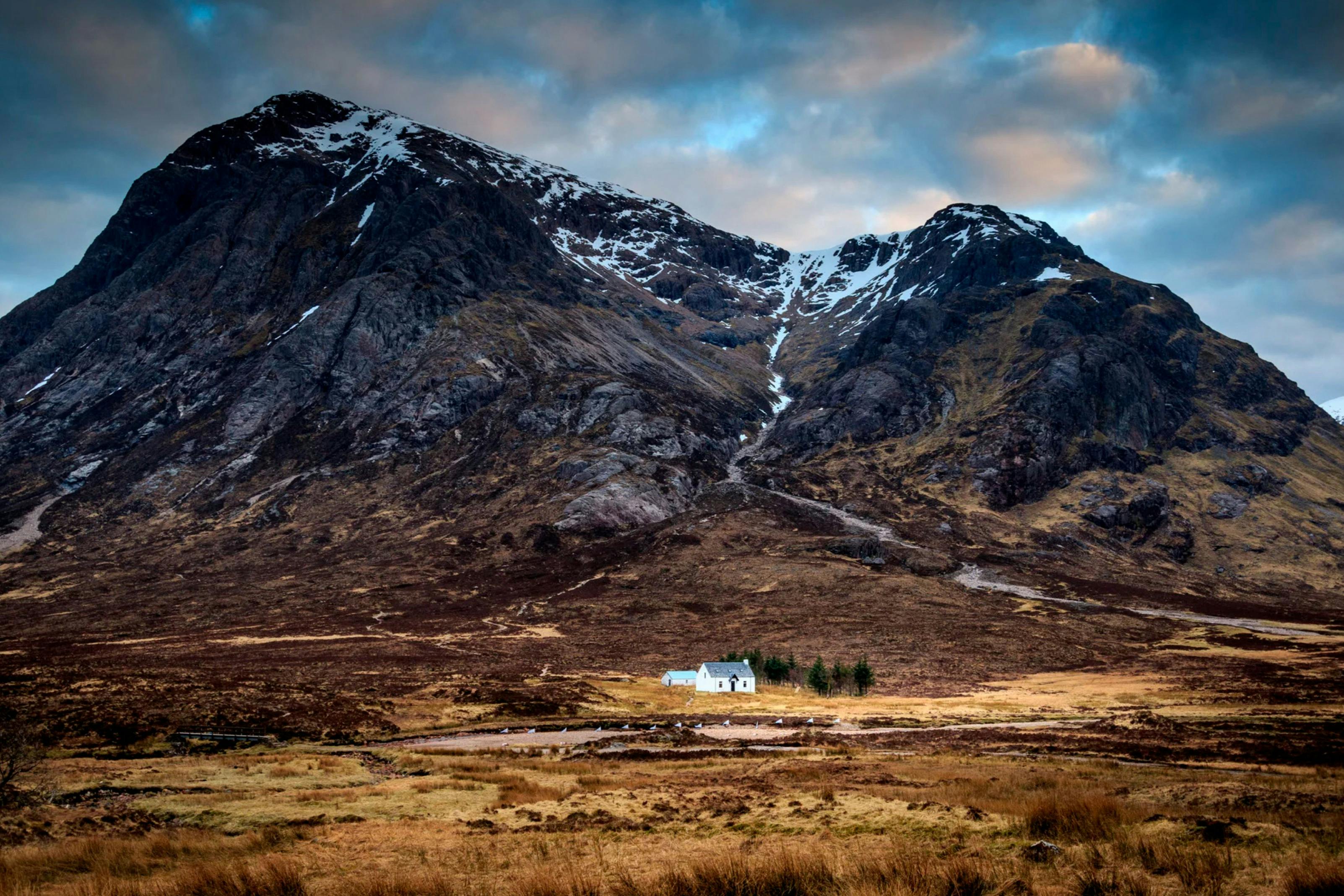 Небольшой белый дом, окруженный деревьями, стоит в обширном открытом ландшафте с бурными горами на заднем плане.