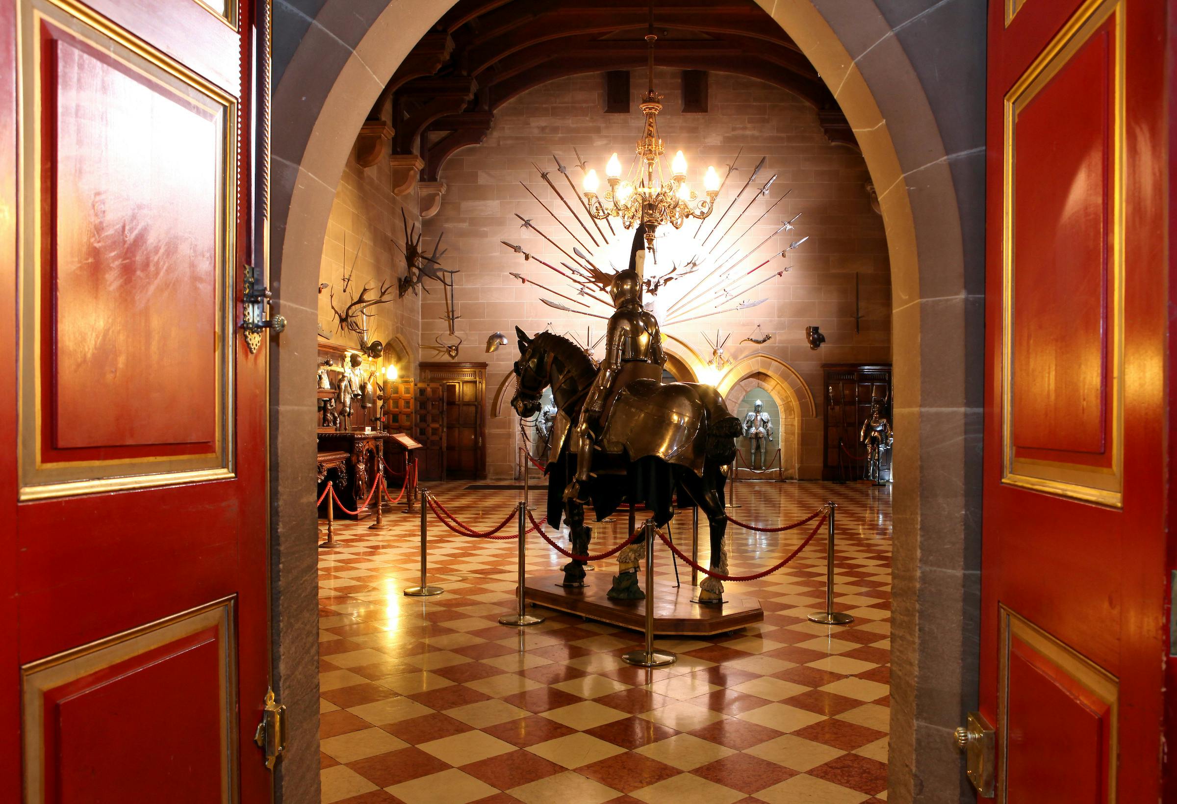A suit of armor on a horse displayed in a historical room with checkered tile floor, chandeliers, and medieval weapons.