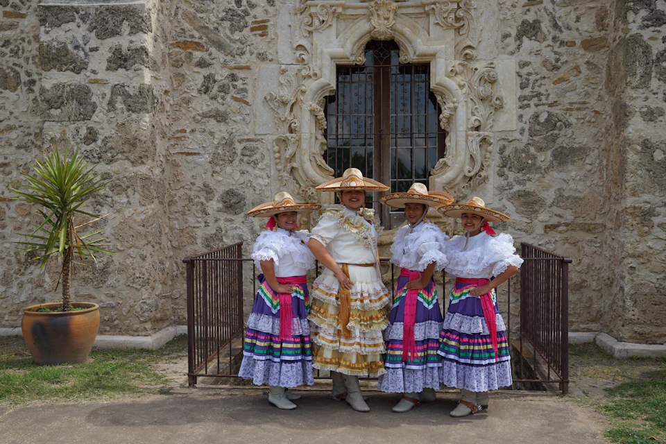 Four women in traditional Mexican dresses and hats stand in front of a historic stone building with ornate details.
