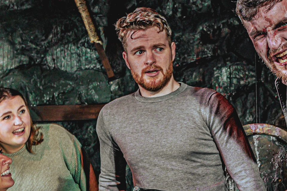 Three people look at a man holding a saw with a grimace in a dimly lit, eerie room decorated with a severed head on a pike.