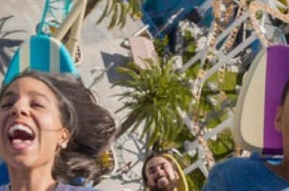 Three people on a rollercoaster, visibly excited and screaming, with the track and park visible in the background.