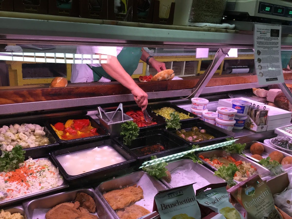 Person preparing a sandwich behind a deli counter displaying various food items like salads, vegetables, and prepared dishes.