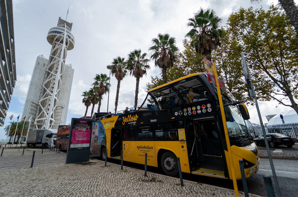 A yellow open-top tour bus parked beside a tall structure and palm trees on a cloudy day.