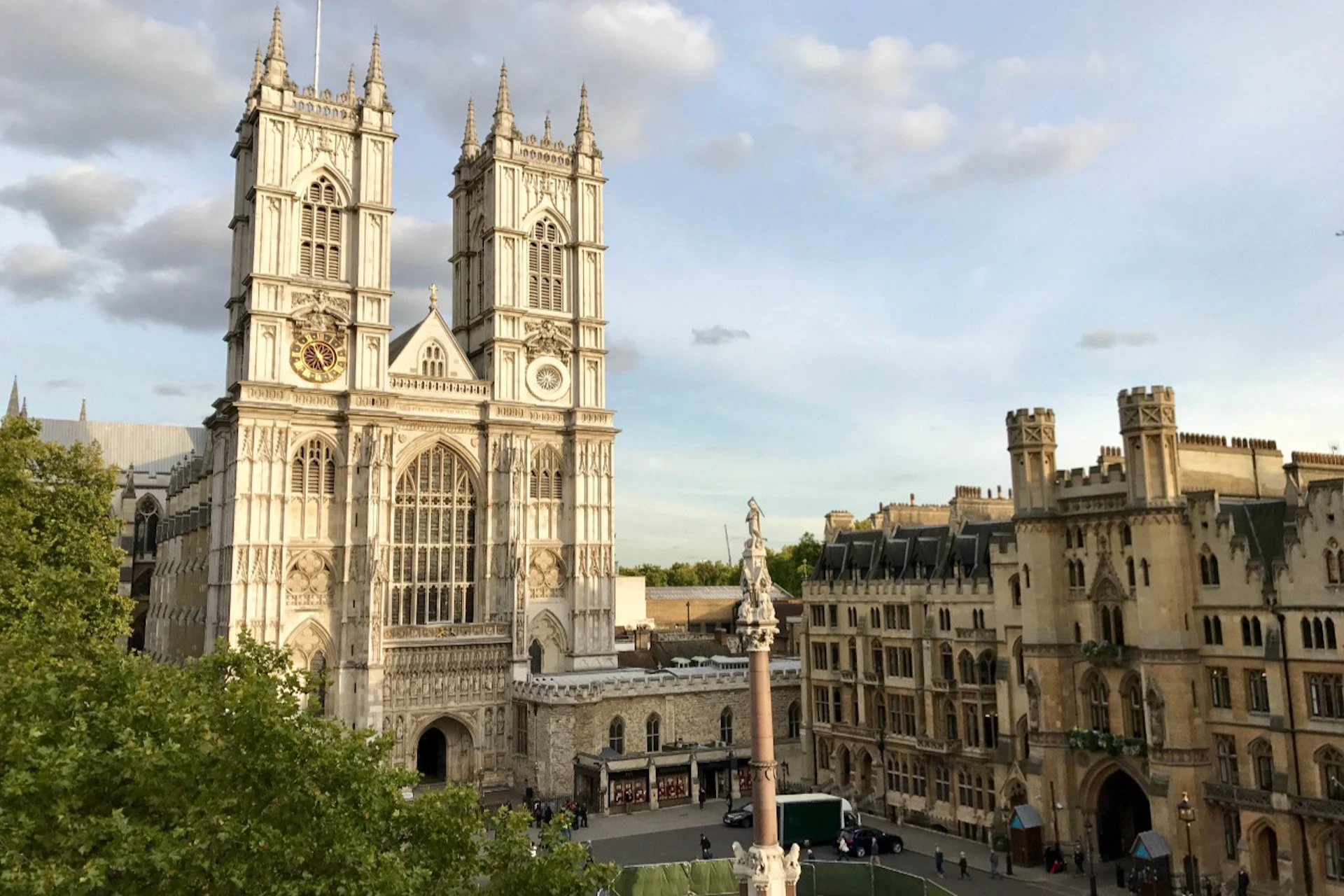 A historic, gothic-style cathedral with twin towers next to traditional buildings, a statue, and a tree, under a partly cloudy sky.