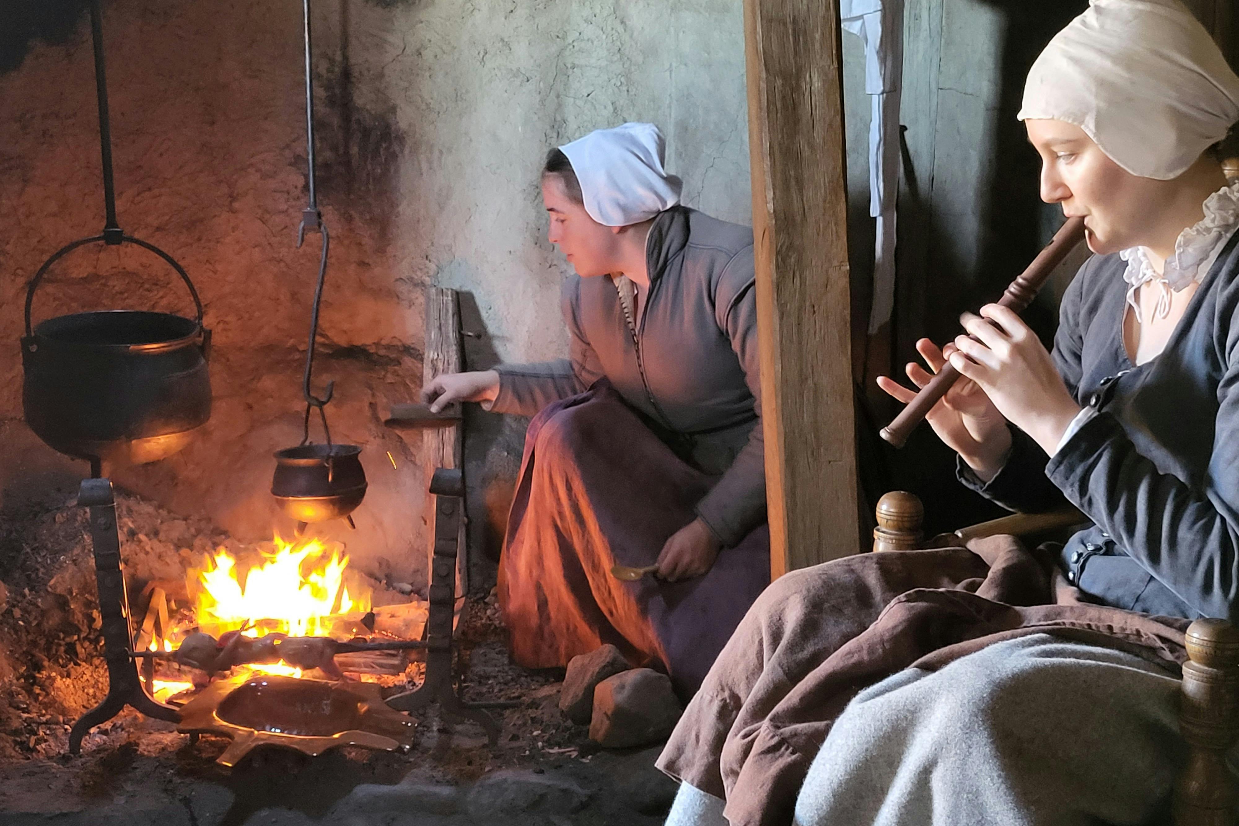 Dvě ženy v historickém oblečení v rustikálním interiéru. Jeden hraje na dřevěnou flétnu, zatímco druhý má sklon ke krbu.
