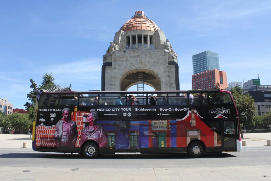 A double-decker tour bus in front of a historic monument under a clear blue sky, with people on the bus's upper deck.
