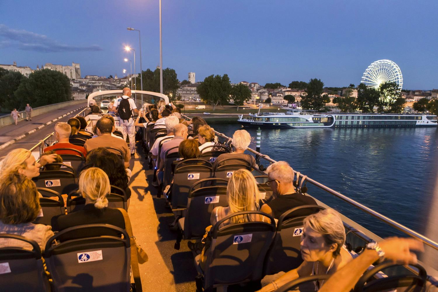 Lidé sedící za soumraku na otevřeném autobusu přecházejícím most s výhledem na řeku s velkým ruským kolem a budovami v pozadí.