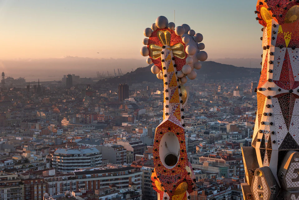 A colorful, ornate spire adorned with mosaic tiles towers over a densely built cityscape at sunset.