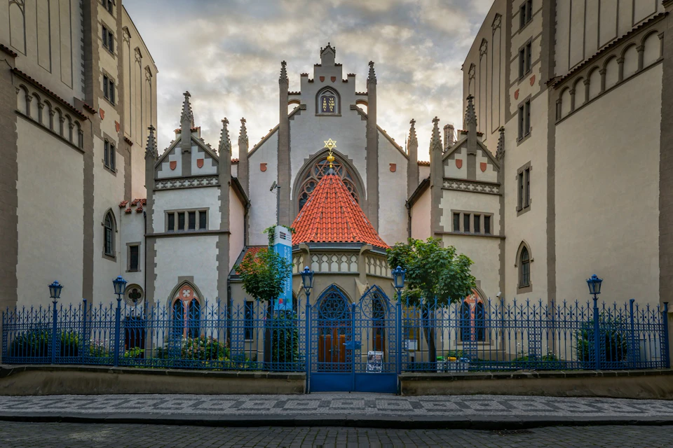 Historic building with ornate facade, arched windows, and a red-tiled roof behind a blue wrought-iron fence and gate.