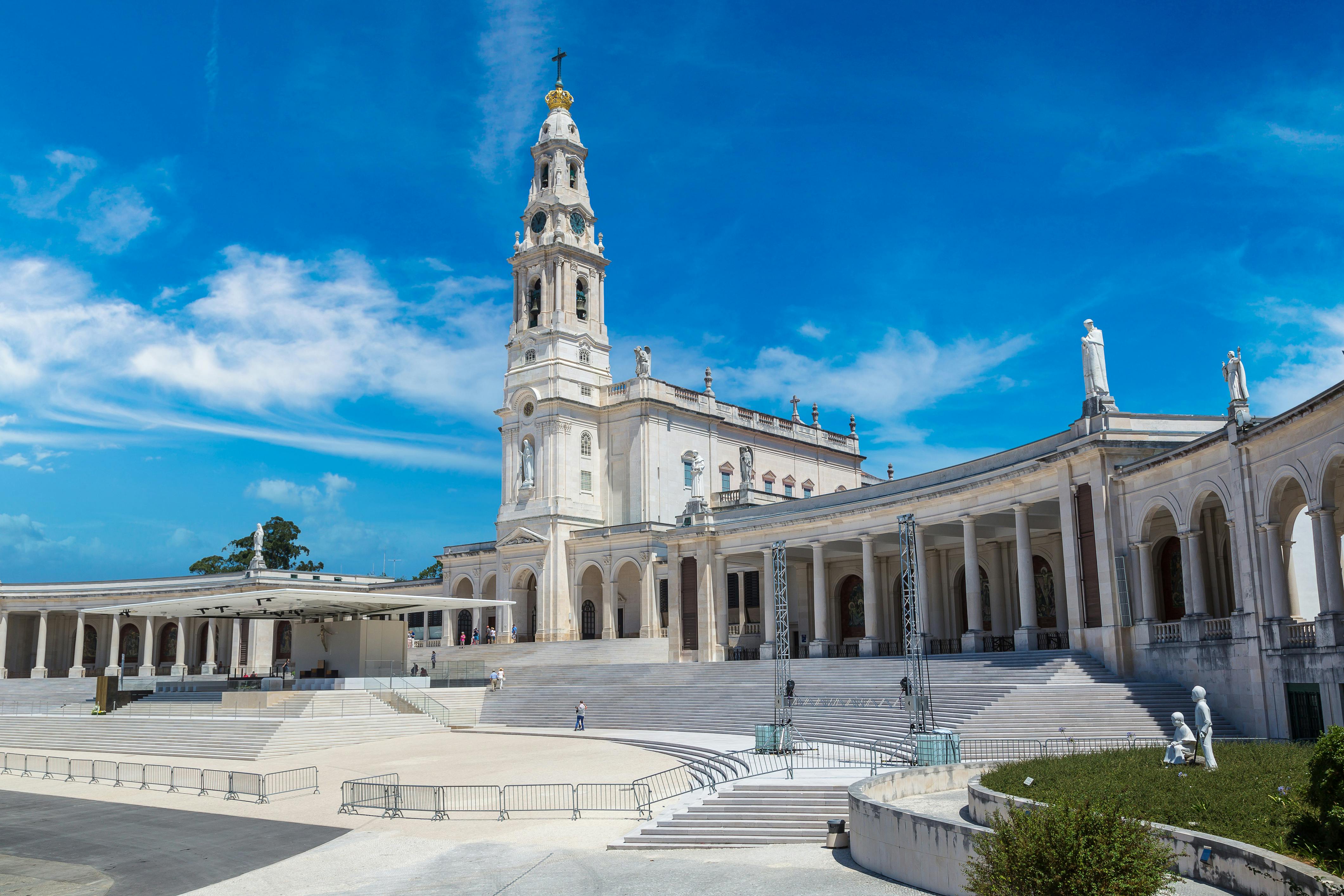 A large white basilica with a tall bell tower and colonnades, under a bright blue sky. Few people are scattered around the foreground.