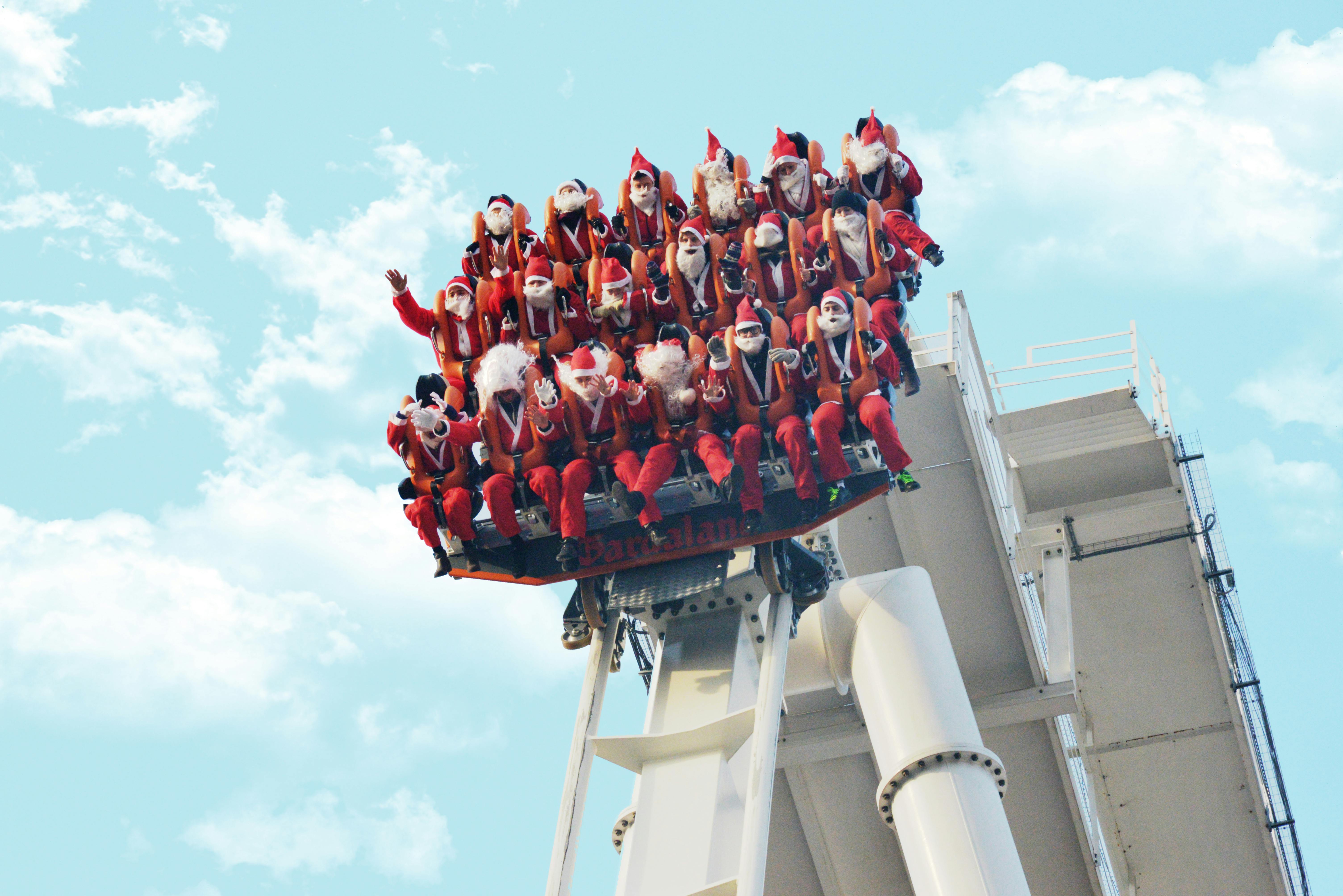 Skupina lidí oblečených jako Santa Claus jezdí na zábavní jízdě na padací věži proti jasně modré obloze.