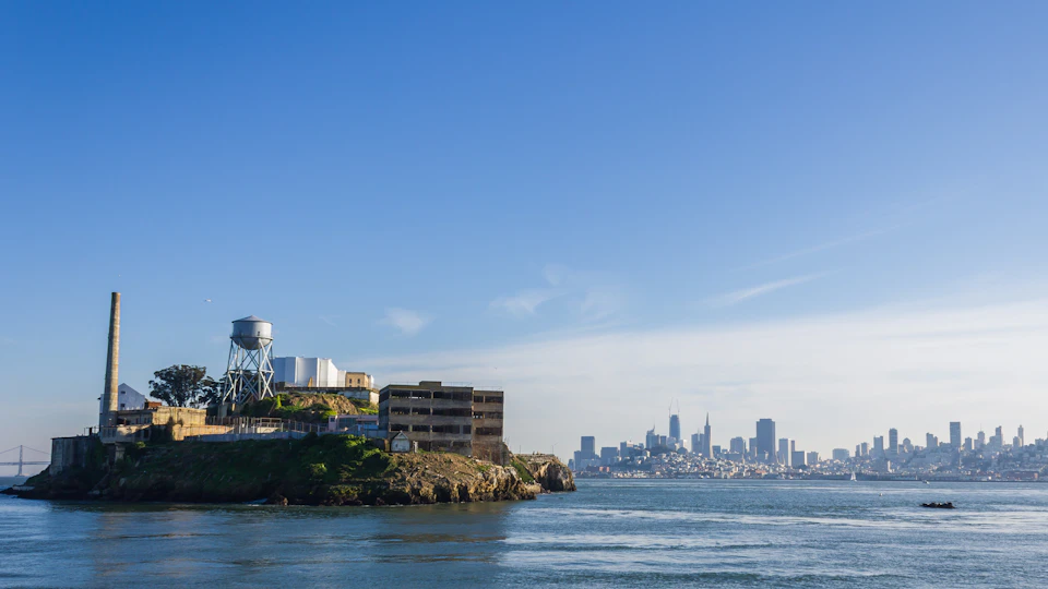 A dilapidated building and water tower on an island foreground, with a city skyline and blue sky in the background.