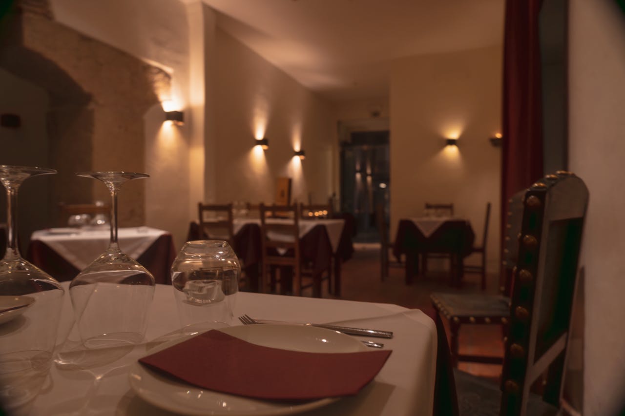 前景にはきれいにセットされたテーブルがあり、バックグラウンドにはいくつかの空の屋根付きテーブルがある、薄暗い照明のレストラン。