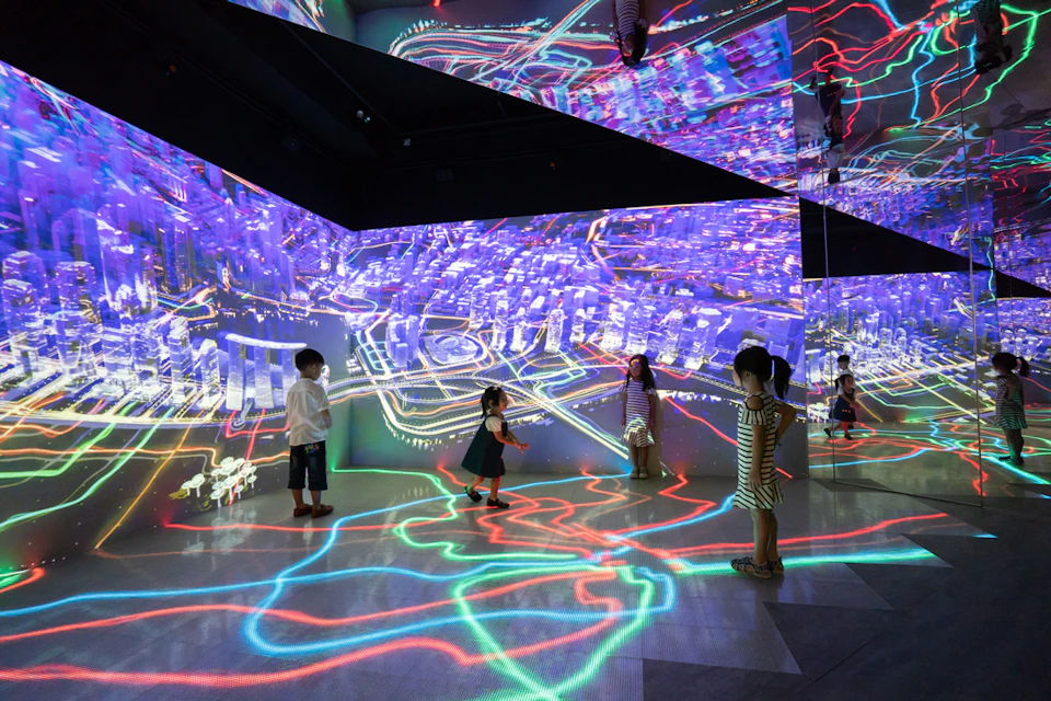 Children exploring a brightly lit, immersive digital art exhibit featuring colorful, abstract cityscape projections on walls and floor.