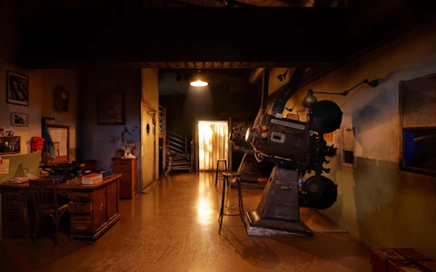 Habitación tenuemente iluminada con un proyector de cine antiguo, un escritorio de madera y una puerta resplandeciente. Escalera de caracol visible al fondo.