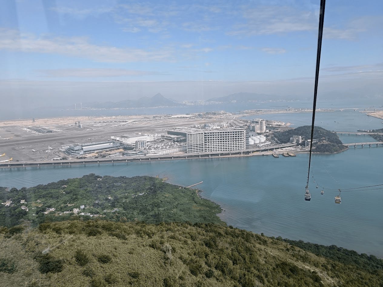Aerial view of a coastal urban area with a large building, water, greenery, and cable cars suspended over the scene.