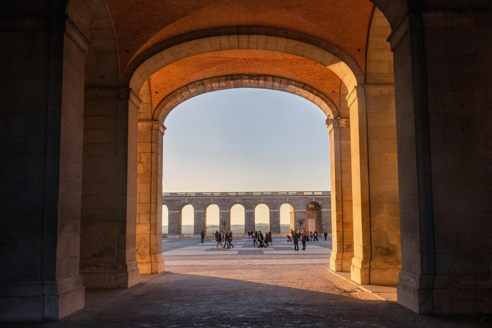 People walking through a large stone archway, with an open courtyard and a series of arches in the background at sunset.