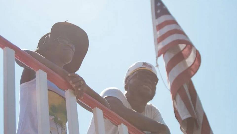 Two people lean on a railing outdoors, one wearing a wide-brimmed hat and the other a captain's hat, with an American flag behind them.