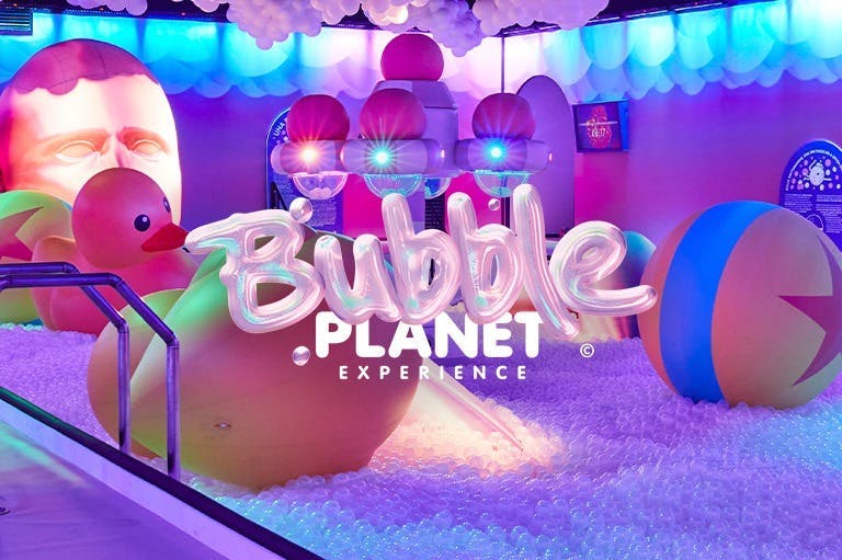 Kolorowy wewnętrzny plac zabaw z dużymi gumowymi kaczuszkami, piłkami plażowymi i tabliczką „Bubble Planet Experience” pośród oświetlonych dekoracji.