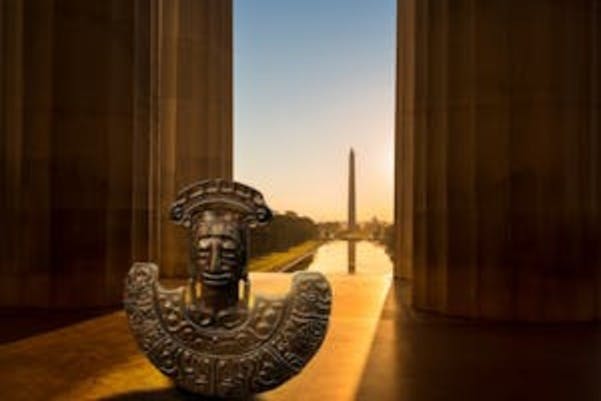 Ornate sculpture of a face and headdress between large columns, with the Washington Monument and Reflecting Pool in the background at sunset.