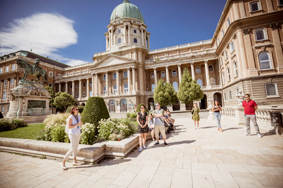 People walking and sitting in front of a historic building with a dome, surrounded by trees and gardens.