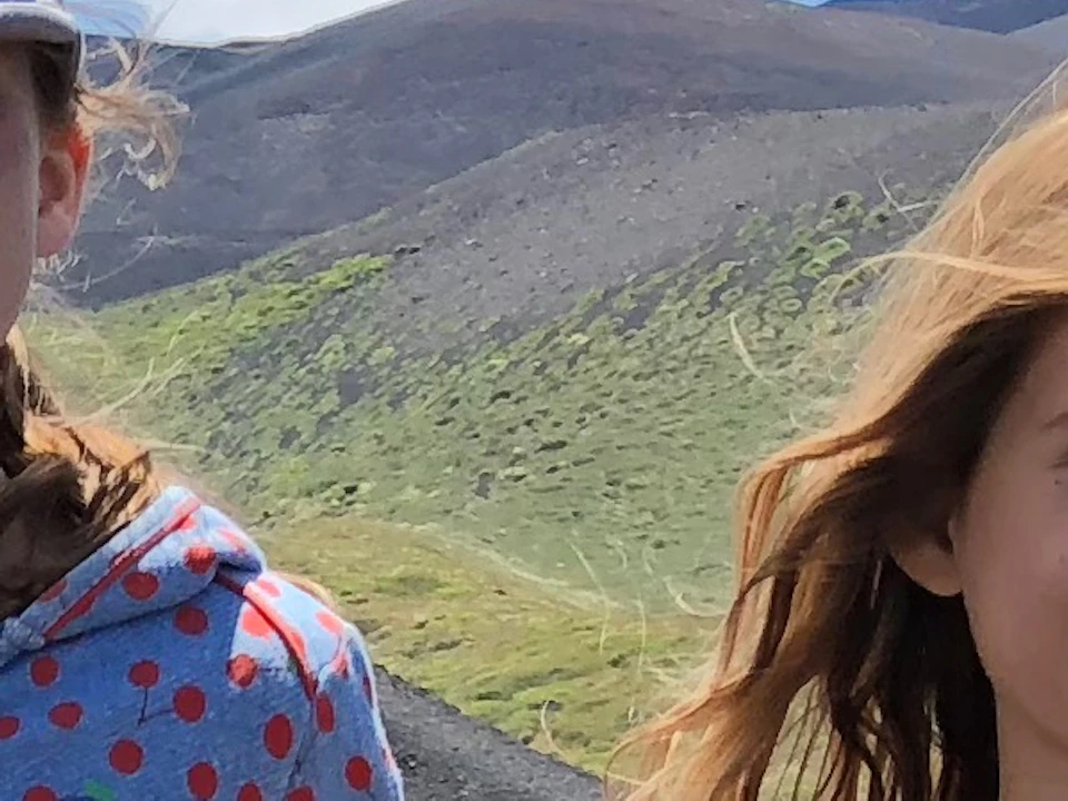 Two smiling girls in casual clothing, standing on a rocky terrain with mountains in the background under a bright blue sky.