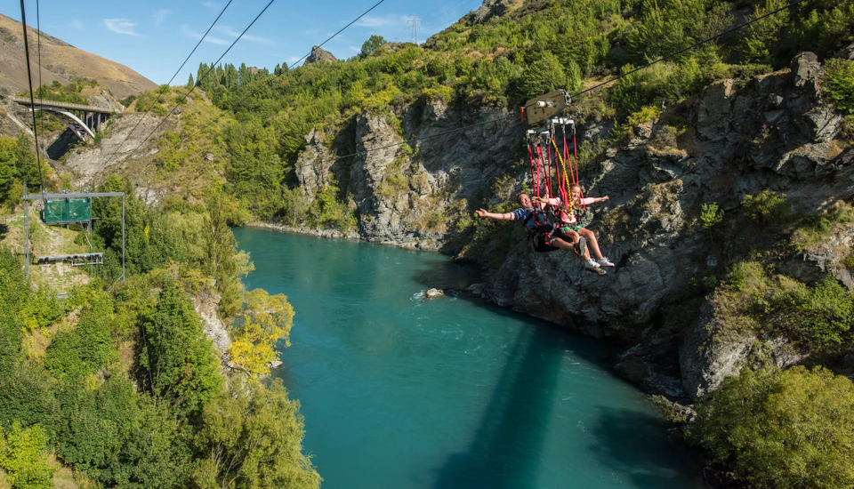Two people riding a zip line above a turquoise river running through a forested and rocky canyon under a clear sky.