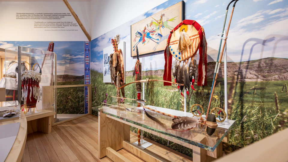 Museum display featuring Native American artifacts, including feathered headdresses, a bow, and traditional artwork on a backdrop of grasslands.