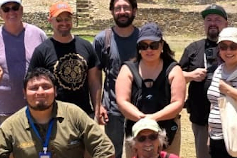 A group of people posing together outdoors in a sunny area with trees and ancient stone steps in the background.