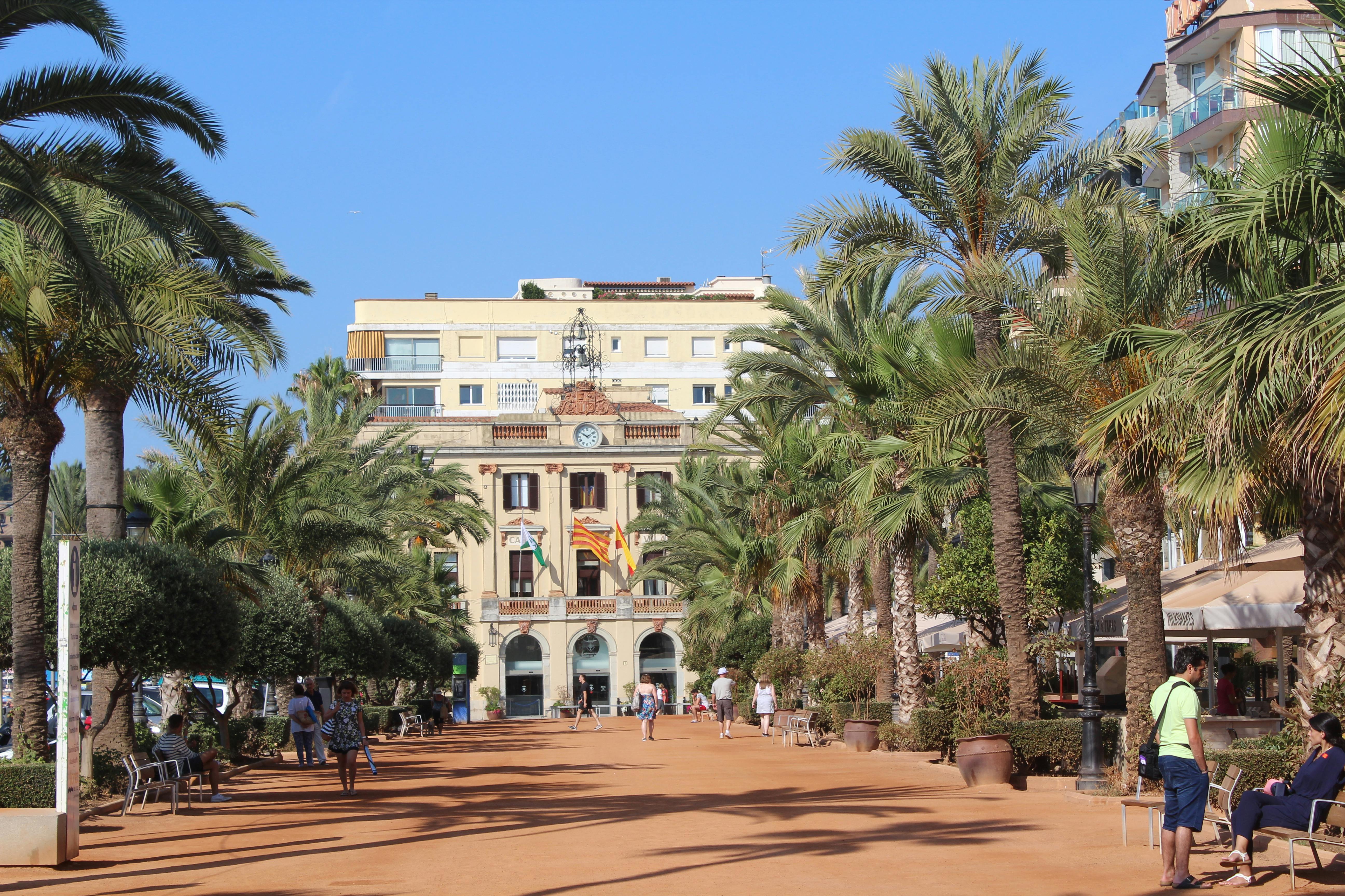 在晴朗的日子里，人们沿着绿树成荫的小径走向一座带有旗帜、时钟和拱门的历史建筑。