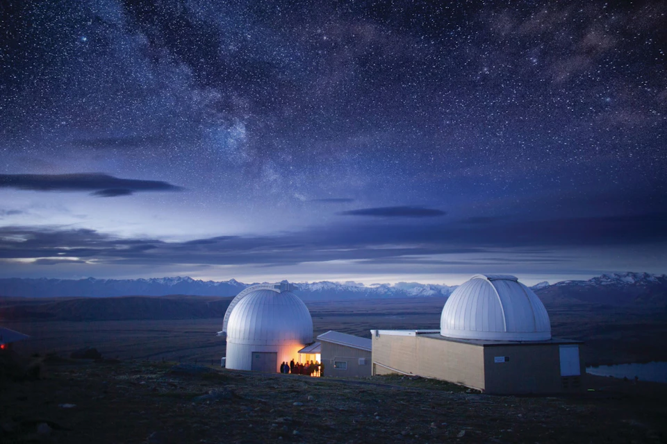 Two observatory domes under a starry night sky with silhouetted mountains in the background and a lit entrance visible.