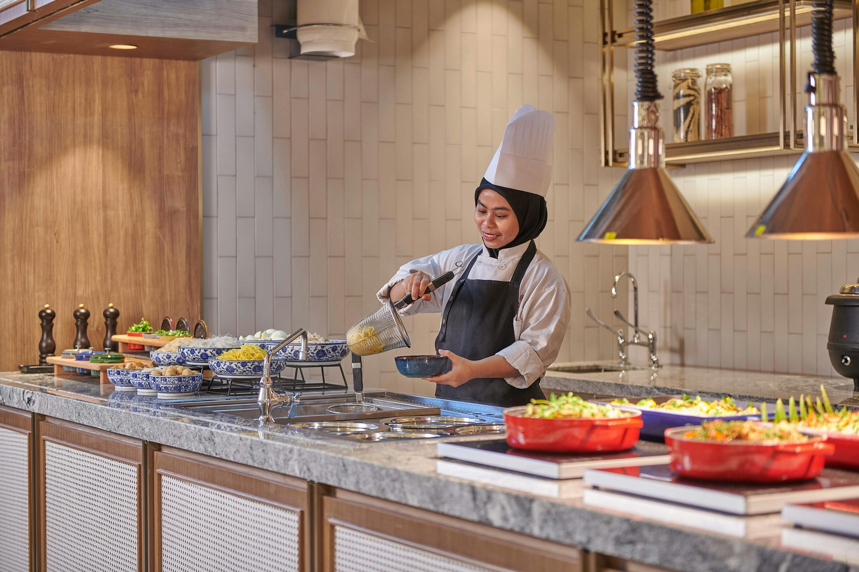 A chef in a white uniform and hat serves food from a buffet counter in a well-lit modern kitchen setting.