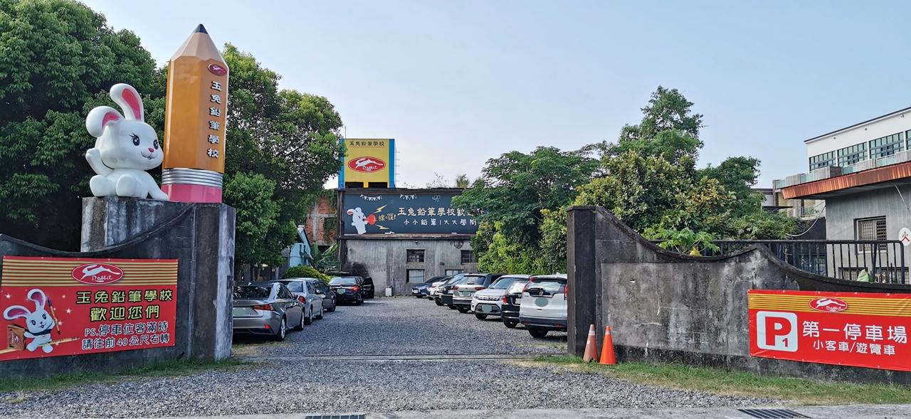 A parking lot with several cars parked on gravel, surrounded by trees, and a building with signs in the background.
