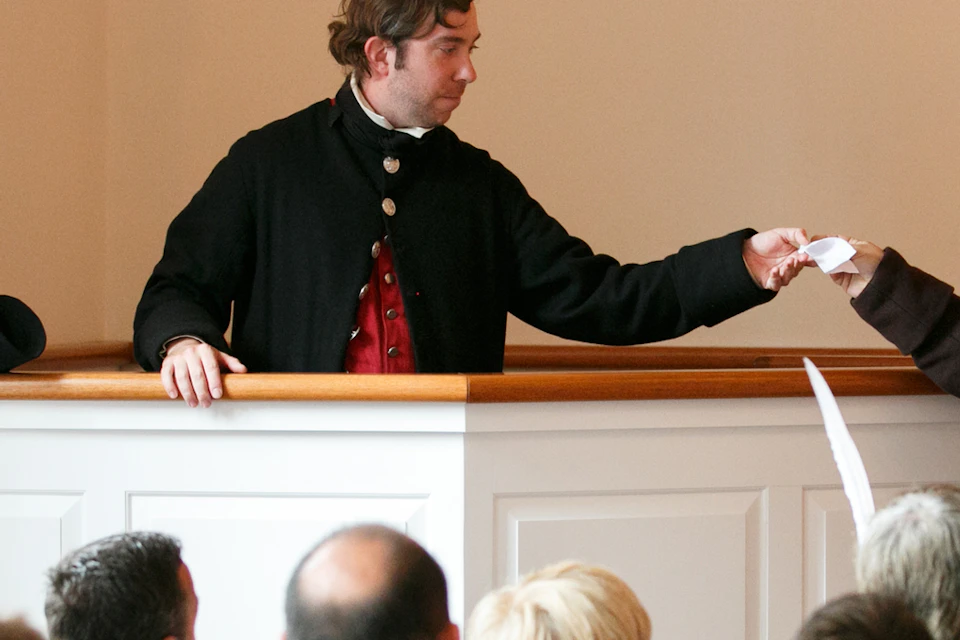 A man in colonial attire speaks from a pulpit to an audience, while another costumed man hands him a document.