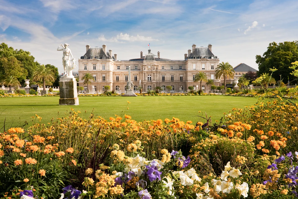 Ornate building with a French flag, surrounded by a garden with colorful flowers, a grass lawn, and a statue in the foreground.