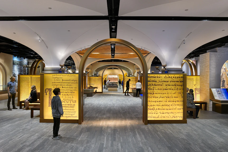 Modern museum exhibit hall with illuminated ancient manuscript displays and several visitors walking through arched doorways.