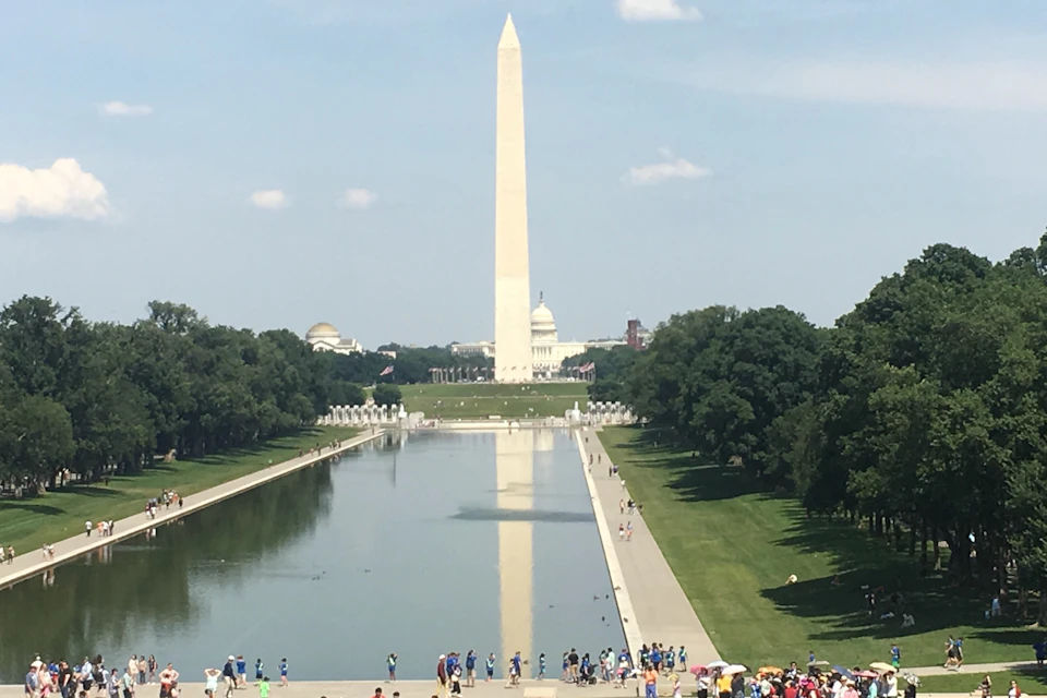 The Washington Monument reflected in the Reflecting Pool, with the U.S. Capitol in the background and visitors nearby.