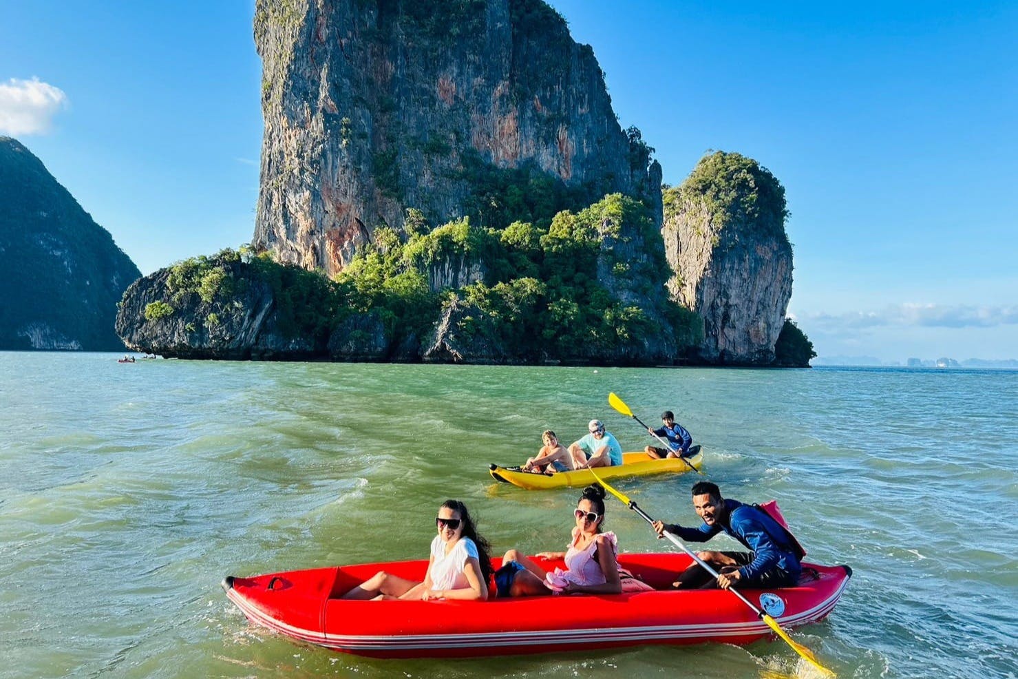 Lidé jezdí na kajaku poblíž svěžích, skalnatých útesů na čisté, otevřené vodě pod modrou oblohou.