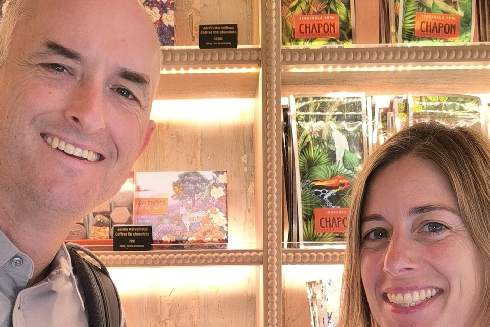 Two people smiling in front of a display shelf filled with packaged goods. One holds a chocolate dessert.