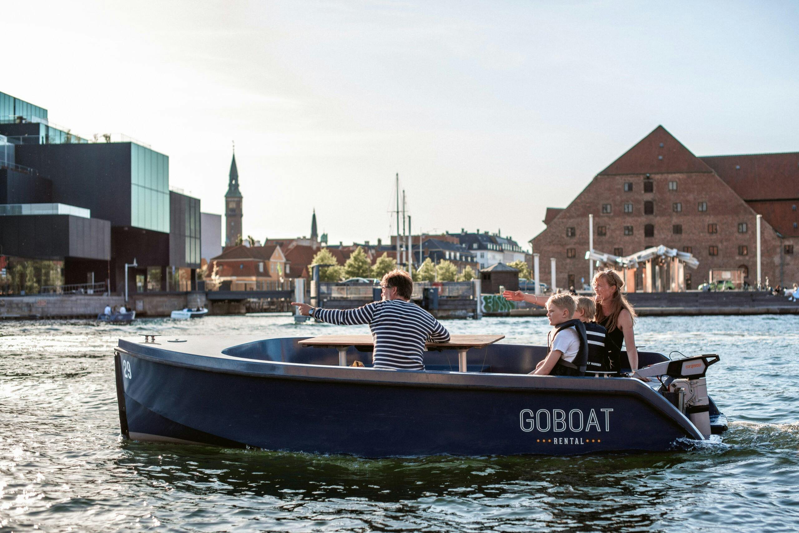 Three people in a rental boat on a river with a large brick building and other structures in the background under a clear sky.