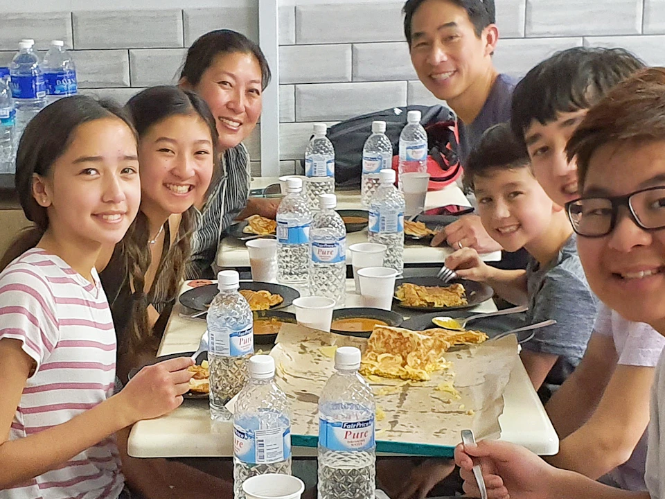 Group of people smiling at a dining table with plates of food, water bottles, and bowls of soup.