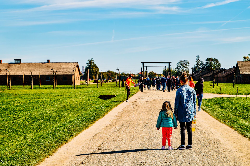 A woman and a child walk on a path towards a group of people near brick buildings and green grass under a clear blue sky.