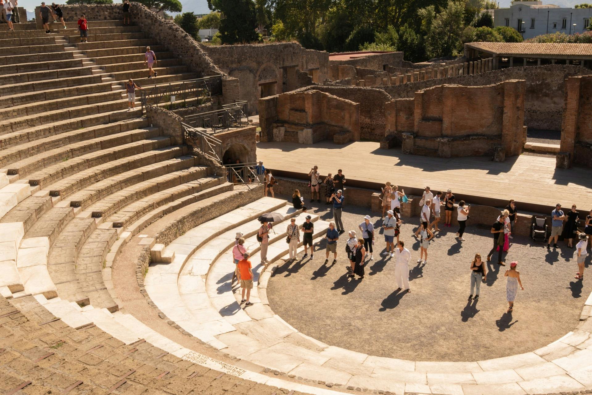 遺跡や緑に囲まれた、晴れた空の下に階層的な石の座席がある古代の円形劇場を観光客のグループが探索しています。
