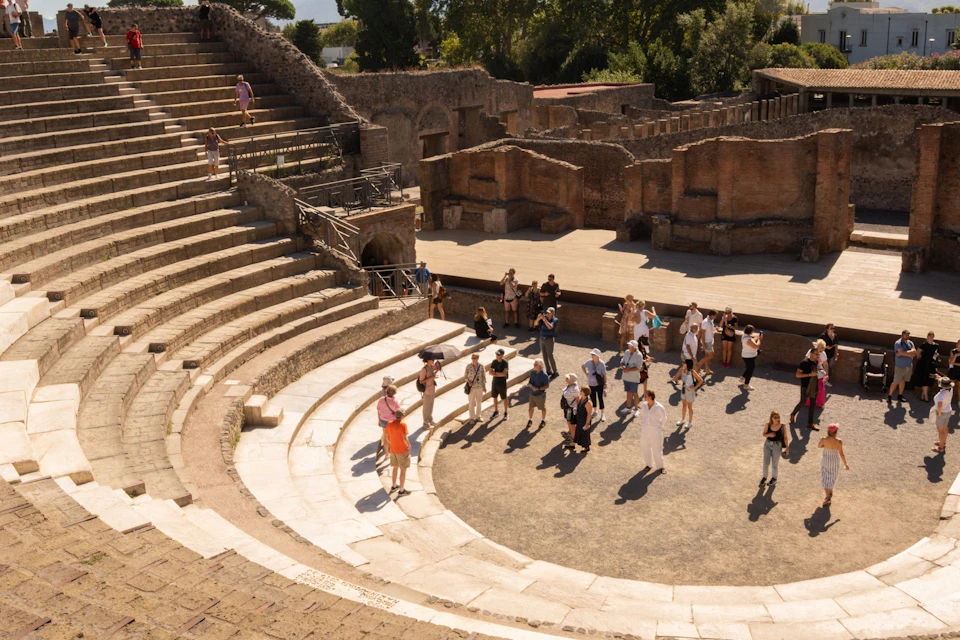 A group of tourists explores an ancient amphitheater with tiered stone seating under a sunny sky, surrounded by ruins and greenery.