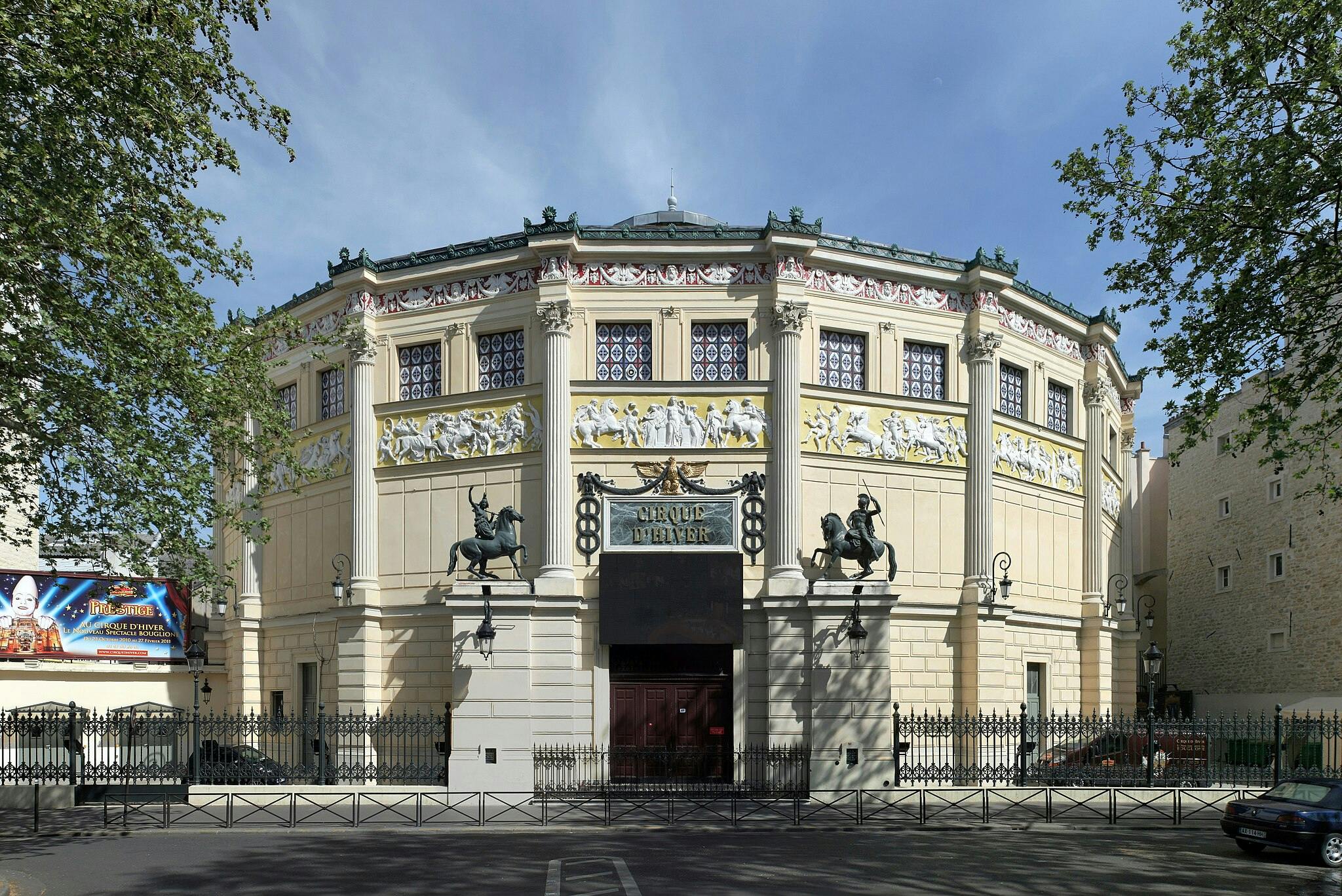입구에 고전적인 기둥, 화려한 동상, 'Cirque d' Hiver '간판이 있는 크림색 팔각형 건물. 나무 프레임 측면.