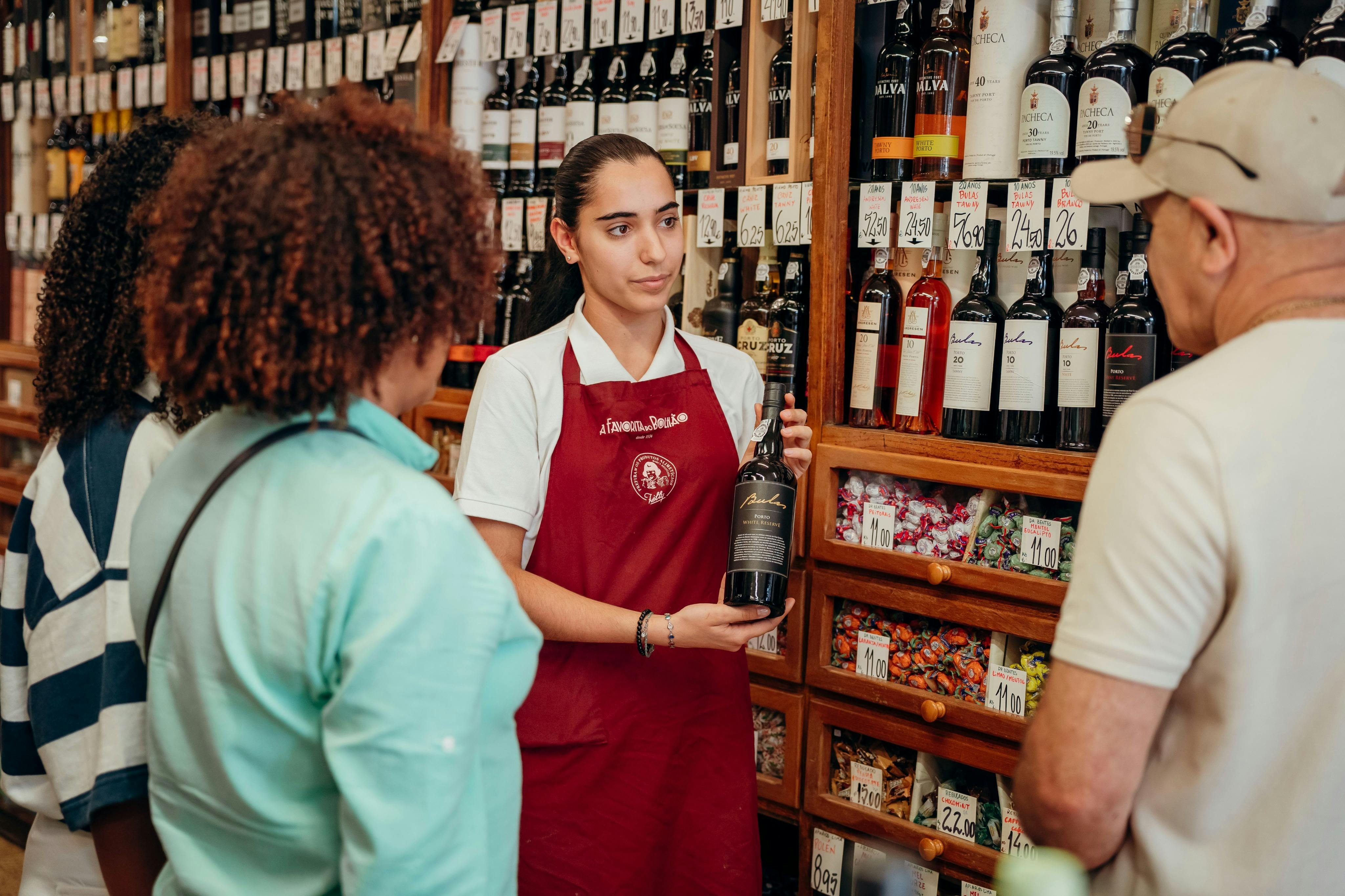 Eine Verkäuferin in einer roten Schürze hält eine Weinflasche, während sie mit zwei Kunden spricht. Im Hintergrund befinden sich mit Weinflaschen gefüllte Regale.