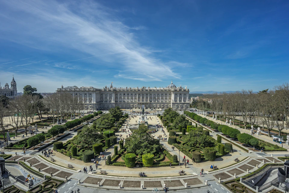 A large palace with a symmetrical garden in front, featuring trees, paths, and people walking under a clear blue sky.