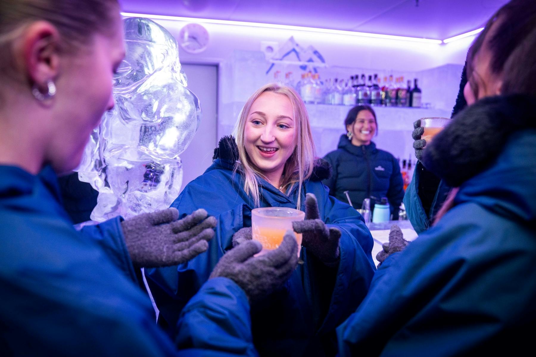 겨울 재킷을 입은 사람들은 얼음 조각과 웃는 바텐더를 배경으로 얼음으로 만든 바에서 음료를 즐깁니다.