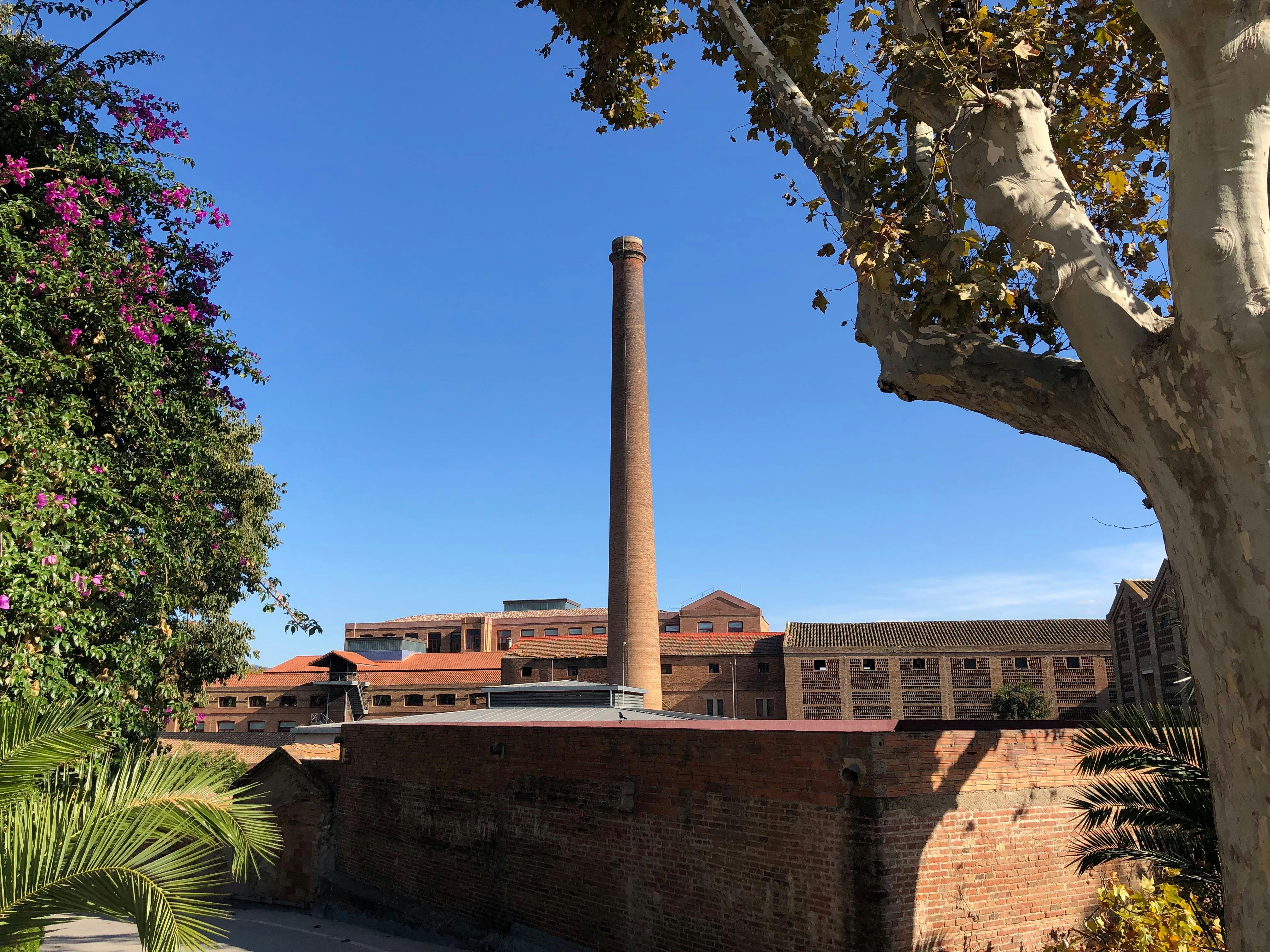 Tall brick chimney amidst red brick buildings, surrounded by trees and clear blue sky.